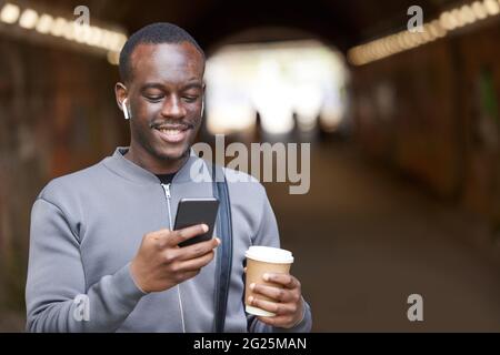 Lächelnder Junger Mann In Der Stadt Überprüft Soziale Medien Und Nachrichten Auf Dem Mobiltelefon, Während Er Schnurlose Ohrhörer Trägt Stockfoto