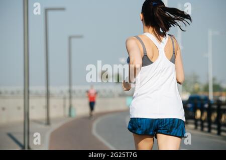 Friedliche, athletische Frau, die draußen auf der neuen Strecke läuft. An einem sonnigen Tag unter einem klaren blauen Himmel. Sie trägt ein weißes ärmelloses Hemd und eine Mini-Shorts Stockfoto