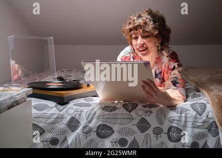 Junge Frau, die auf ihrem Bett lag, eine Schallplatte hörte und auf den Ärmel schaute Stockfoto