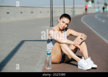 Porträt einer ruhenden athletischen Frau in einer Sportkleidung, die draußen auf der Strecke sitzt und mit einem subtilen falschen Lächeln auf die Kamera blickt. An einem sonnigen Tag und Stockfoto