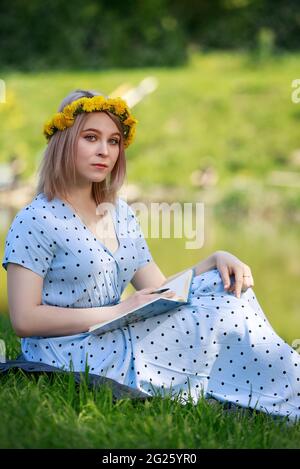 Schöne blonde junge Frau, die für ihre Prüfungen studiert, sitzt auf einem Gras in der Nähe des Sees und liest in einem hellblauen Kleid ein Buch. Warm. Sommer. Im Freien. Stockfoto