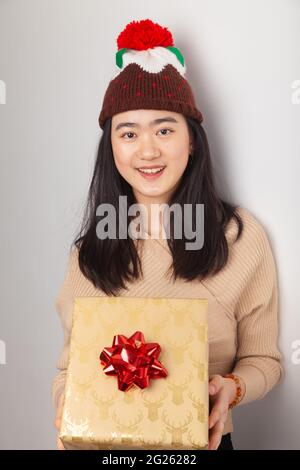 Eine asiatische Frau, die einen weihnachtlichen Puddinghut trägt und ein Weihnachtsgeschenk hält. Stockfoto