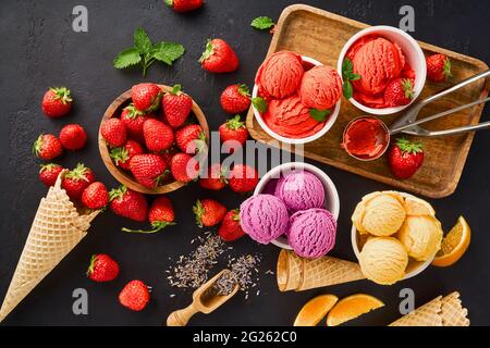 Haufen von Erdbeere und Orange, getrockneter Lavendel mit Tassen Eis und Zapfen. Lavendel, Erdbeere und Orangensorbet auf schwarzem Hintergrund. Stockfoto