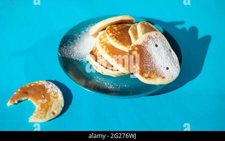 Pfannkuchen werden mit Puderzucker auf einem türkisfarbenen Teller bestreut. Gebissen von Pfannkuchen. Platz für Text kopieren. Blauer Hintergrund. Stockfoto