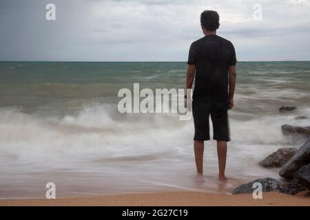 Ein Mann steht an einem Strand mit Wasser, das um seine Füße spritzt, Bewegungsunschärfen bei langer Belichtung, konzeptuellem Bild mit Stimmung und Umgebung. Stockfoto