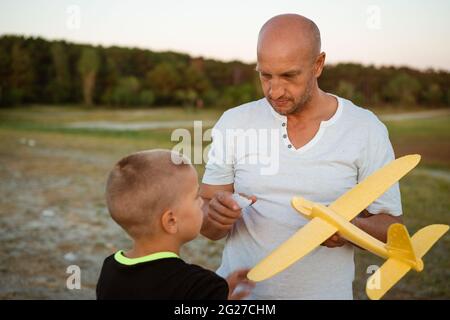 Weicher Fokus von Vater und Sohn spielen Spielzeug Flugzeug in der Wiese bei Sonnenuntergang mit glücklichen Emotionen. Familien-, Urlaubs- und Reisekonzept. Bei Sonnenuntergang im Sommer starten sie ein Flugzeug vor dem Hintergrund des Himmels Stockfoto