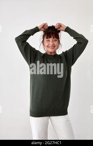Lächelndes weibliches Model mittleren Alters in legerem, bequemen Wollpullover und weißen Hosen, die die Hände heben und die Kamera betrachten Stockfoto