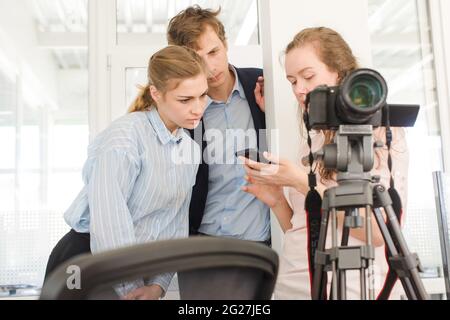 Eine Gruppe junger kreativer Mitarbeiter, die mit einer professionellen Kamera in der Nähe eines Stativs stehen und Fotos auf dem Mobiltelefon diskutieren, während sie in der modernen Umgebung zusammenarbeiten Stockfoto