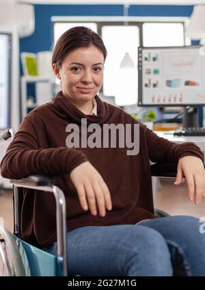 Nahaufnahme der gelähmten behinderten behinderten Frau Arbeitskollegin, die lächelnd auf die Kamera schaut, die im Wirtschaftsbüro am Computer mit Daten arbeitet und finanzielle Informationen prossesst. Stockfoto