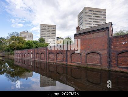 Reihe aus Wohnhäusern aus den 1960er und 1970er Jahren mit Hochhäusern und Wolkenkratzern hinter roten Backsteinwänden, die in den Wasserstraßen des Birmingham-Kanals spiegeln. Stockfoto