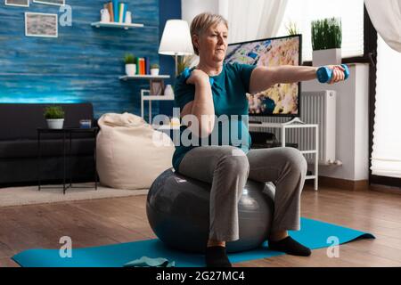 Sportlicher Rentner in Sportswear, der online Aerobic-Workout mit einem Tablet auf schweizer Ball beobachtet und dabei Körperwiderstand trainiert. Ältere Frau führt im Wohnzimmer mit Hanteln eine Armübung durch Stockfoto