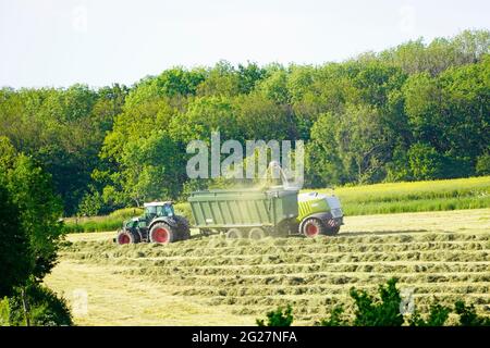 Zwei landwirtschaftliche Fahrzeuge ernten Heu. Sammeln von Heu für Silage. Hügelige grüne Landschaft. Stockfoto