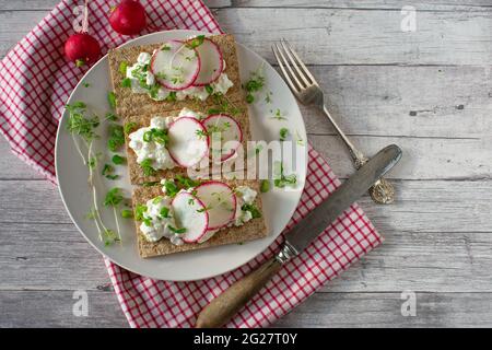 Ein Teller mit Knäckebrot-Sandwiches, gekrönt mit Quark und rotem Rettich. Serviert auf rustikalem und hölzernen Hintergrund auf kariertem Geschirrtuch Stockfoto