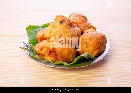 Potato Bonda, ein würziger, indischer Snack, der auf einer Stahlplatte mit Bananenblatt auf traditionelle Weise und isoliert auf einem strukturierten Hintergrund aus Holz serviert wird. Stockfoto