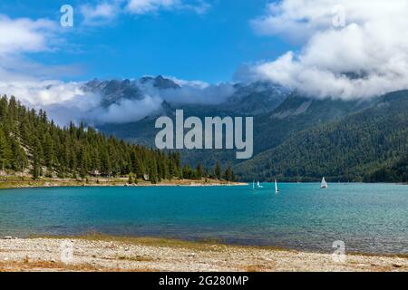 Blick auf das wunderschöne Wasser am Lago di Ceresole und die grünen Bäume an den Berghängen unter dem wehenden Himmel mit weißen Wolken im Piemont, Norditalien. Stockfoto