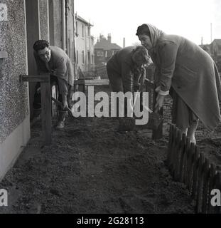 1960er Jahre, historisch, vor einem Reihenhaus, ein Mann und zwei Frauen mit sich verbiegenden Pik, während sie an einem Pfad arbeiten und Boden für die Pflasterplatten legen, Kelty, Fife, Schottland, Großbritannien. In diesem Dorf, das an der Grenze zwischen Fife und Perthshire liegt, standen die Menschen in diesem Dorf vor einer ungewissen wirtschaftlichen Zukunft, da die Kohlebergwerke, Die einst die Mehrheit der lokalen Arbeiter seit 1873 beschäftigt hatte, als die erste Tiefmine, die Lindsey Mine, versenkt wurde, wurden geschlossen. Stockfoto