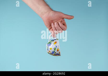 Hand und Tasse auf blauem Hintergrund. Mann hält weiße Tasse mit bemalten Blumen. Keramik Becher Tasse für Tee oder Kaffee hell dekoriert in blau und gelb c Stockfoto