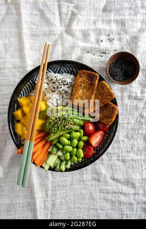 Draufsicht auf eine hausgemachte vegane Poke-Schale mit Tofu, Mango, Avocado, Karotte, Edamame, Tomate, Gurke auf weißem Stoffhintergrund, vertikal Stockfoto