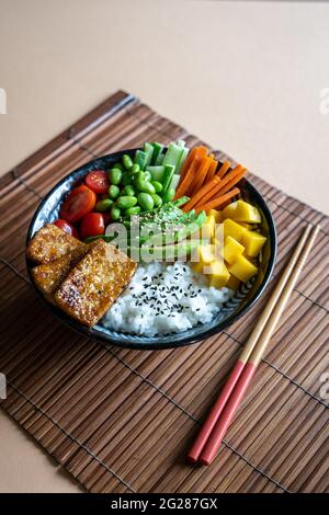 Seitenansicht einer hausgemachten veganen Poke-Schale mit Tofu, Mango, Avocado, Karotte, Edamame, Tomate, Gurke auf einer Mandel- und Bambusmatte Hintergrund, vertikal Stockfoto