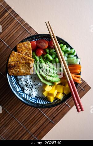 Flatlay einer hausgemachten veganen Poke Schale mit Tofu, Mango, Avocado, Karotte, Edamame, Tomate, Gurke auf einer Mandel- und Bambusmatte Hintergrund, vertikal Stockfoto