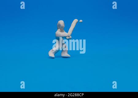 Eine weiße Plastilin-Puppe, die auf blauem Hintergrund Baseballspiele übt. Die Puppe ist ein Teig und hält die Fledermaus bereit, um den Ball zu treffen, der auf ihn zukommt. Stockfoto