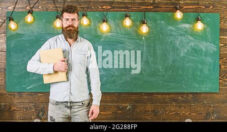 Zurück zum Schulkonzept. Bärtiger Hipster hält ein Buch, eine Tafel auf dem Hintergrund. Mann mit Bart und Schnurrbart im ernsten Gesicht steht vor der Tafel Stockfoto