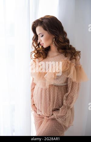 Eine schöne, Schwangere steht in einem beigen Vintage-Kleid neben einem weißen Spitzenschirm und berührt den Bauch mit einem Baby. Stockfoto