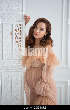 Eine schöne, Schwangere steht in einem beigen Vintage-Kleid neben einem weißen Spitzenschirm und berührt den Bauch mit einem Baby. Stockfoto