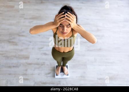 Verärgerte junge Inderin, die über ihr Gewicht unglücklich ist, auf einer Waage stehend, den Kopf enttäuscht haltend, über dem Bild Stockfoto