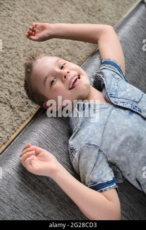 Netter lustiger Junge, der kopfüber auf dem Sofa liegt und die Kamera anschaut, lächelnd verspielter Junge, der zu Hause Spaß hat. Stockfoto