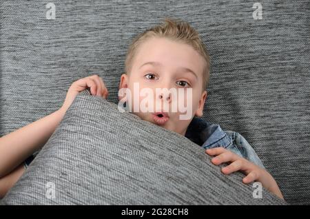 Netter lustiger Junge liegt auf der Couch und macht ein lustiges Gesicht, verspielter Junge, der zu Hause Spaß hat. Stockfoto