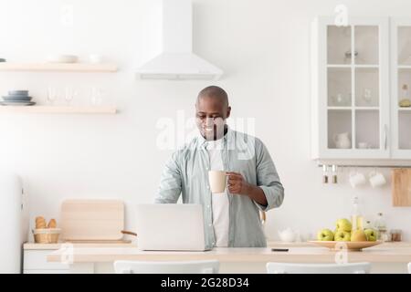 Moderner Arbeiter in Selbstisolation. Glücklicher reifer afroamerikanischer Mann mit Tasse, Kaffee trinken und am Laptop arbeiten Stockfoto