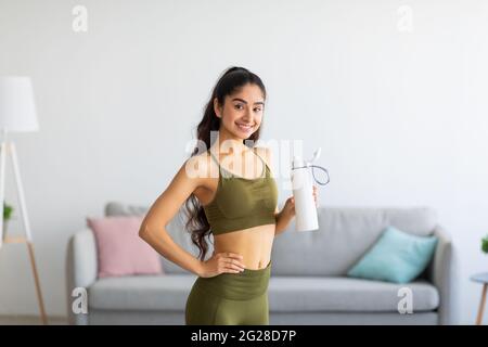 Millennial Indian Frau in Sportkleidung Trinkwasser oder Protein-Shake aus der Flasche zu Hause Stockfoto