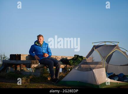 Junger Mann, der eine Tasse Kaffee in der Hand hält und auf einer Holzbank in der Nähe des Touristenzelts sitzt. Männlicher Reisender, der sich im Freien in der Nähe des Zeltlagers mit blauem Himmel auf dem Hintergrund ausruhte. Konzept von Wandern, Reisen und Camping. Stockfoto