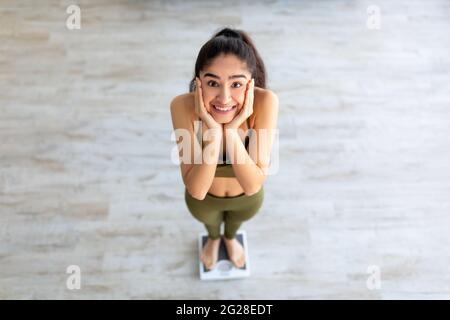 Hübsche indische Frau auf einer Waage, die ihr Gesicht in Aufregung hält und nach erfolgreichem Abnehmen zu Hause, über dem Bild, wiegt Stockfoto