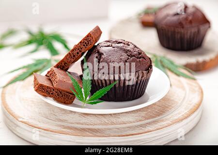 UnkrautMuffins mit Cannabis, Milchschokolade und Cannabisblättern auf weißem Brett auf weißem Tisch serviert. Muffins mit Marihuana-Schokolade und CBD-Gras Stockfoto