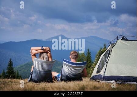Rückansicht der Touristen Paar in der Sonne sonnen, sitzen in Trekkingstühlen in der Nähe des Zeltes auf ihrem Campingplatz und bewundern die umliegende Berglandschaft. Konzept von Wandern, Camping und Beziehungen. Stockfoto