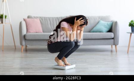 Junge Inderin, die auf einer Waage sitzt, sich über die Gewichtszunahme zu Hause aufregt, Platz kopiert Stockfoto