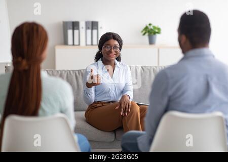 Erfolgreiche Therapie. Professionelle weibliche Psychologin, die den Daumen nach oben gestikuliert, während sie mit einem jungen schwarzen Paar im Büro arbeitete Stockfoto