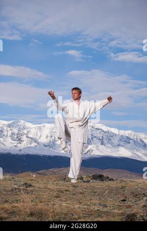 Wushu Master in einer weißen Sportuniform Training auf dem Hügel. Kungfu-Champion trainiert maritiale Künste in der Natur vor dem Hintergrund verschneiter Berge. Stockfoto