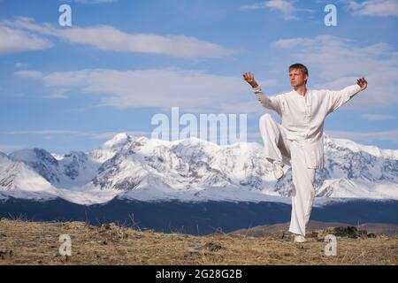 Wushu Master in einer weißen Sportuniform Training auf dem Hügel. Kungfu-Champion trainiert maritiale Künste in der Natur vor dem Hintergrund verschneiter Berge. Stockfoto