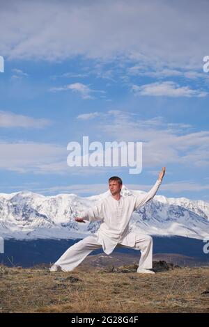 Wushu Master in einer weißen Sportuniform Training auf dem Hügel. Kungfu-Champion trainiert maritiale Künste in der Natur vor dem Hintergrund verschneiter Berge. Stockfoto