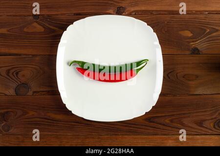 Frische rote und grüne scharfe Chilischoten liegen auf einem weißen original schönen quadratischen Teller auf einem braunen Tisch mit sichtbarer Holzstruktur. Stockfoto