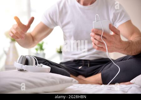 Mann, der ein mit weißen Kopfhörern verbundenes Mobiltelefon mit gekreuzten Beinen auf dem Bett hält Stockfoto
