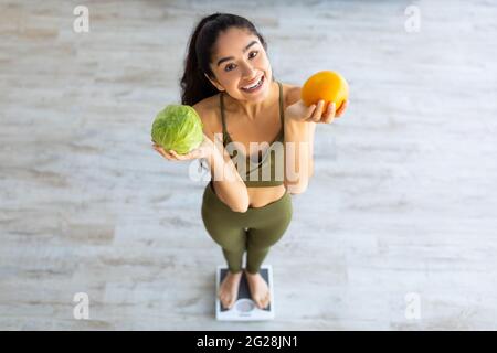 Gesunde Ernährung zur Gewichtsabnahme. Oberhalb der Ansicht der tausendjährigen indischen Dame, die Zitrusfrüchte und Gemüse hält, während sie Schuppen verwendet Stockfoto