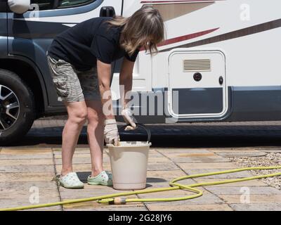 Eine Dame, die ein Wohnmobil besitzt, hält einen Eimergriff und reinigt ihr Freizeitfahrzeug mit einem Schwamm. Sie trägt an einem sonnigen Tag Shorts - Stockfoto