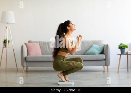 Die ganze Länge der aufgeregt indischen Frau sitzt auf einer Waage zu Hause, überglücklich über den Erfolg ihrer Schlankheits-Diät, Seitenansicht Stockfoto