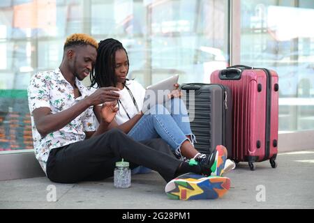 Das junge schwarze Paar kauft die Tickets auf einem Tablet und sitzt mit ihren Koffern am Bahnhof. Urlaubskonzept. Stockfoto