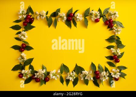Ein einfacher Rahmen aus Blättern, Zweigen, Blumen und Kirschen. Minimale flache Verlegung. Gelber Hintergrund. Stockfoto