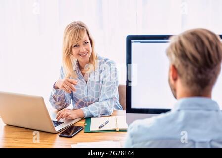 Aufnahme einer glücklichen Geschäftsfrau, die am Laptop arbeitet, während ihre Kollegin ihr gegenüber sitzt und über das Geschäft spricht. Teamarbeit. Stockfoto